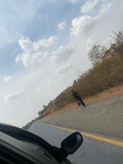 The image presents an outdoor scene, with a diagonal orientation, captured from inside a vehicle, evidenced by the dark car window frame and side mirror visible in the lower left foreground. The location is in the city of Garaku, Nigeria. The upper two-thirds of the frame consists of a light blue sky populated by scattered white cumulus clouds. A two-lane asphalt road, featuring a continuous yellow line on its right shoulder, extends into the distance. To the right of the road, an elevated bank is covered in dry, light-brown scrub vegetation and includes a few trees with green foliage. A single male individual is visible walking along the right shoulder of the road, moving away from the camera's perspective. He is wearing dark attire, consisting of a dark top and dark trousers. He carries a dark, bag-like object in his left hand, held low beside his body.