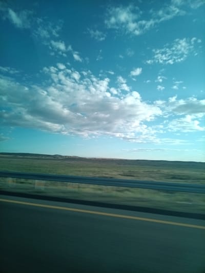 The image depicts a vast, open landscape under a bright, partly cloudy sky. The foreground is dominated by a dark road with a thin yellow line marking its edge, suggesting movement or travel. A metal guardrail runs parallel to the road, separating it from a grassy plain. The plain stretches towards a distant horizon, characterized by rolling hills and muted green and brown tones. Above the land, the sky is a vibrant blue, interspersed with fluffy white clouds of varying sizes and densities. Some clouds are light and scattered, while others are larger and more textured, casting subtle shadows on the land below. The overall impression is one of a clear, possibly late afternoon or early morning, with a sense of spaciousness and serenity. There is no visible text in the image. The scene evokes a feeling of a road trip through a sparsely populated, natural environment.
