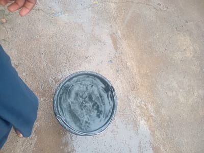 In Girei, Nigeria, a black plastic bucket filled with water is placed on a wet, concrete surface. A person's left leg, clad in dark blue fabric, is visible on the left side of the frame, with their hand also partially visible in the upper left corner. The concrete is damp and appears to have some scattered debris. The water in the bucket is rippling, suggesting recent disturbance or ongoing activity.