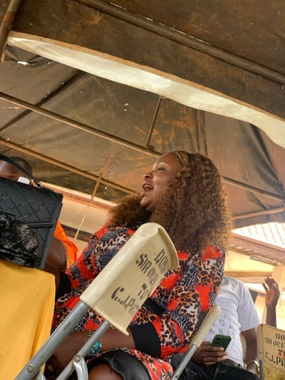 The image captures an outdoor gathering in Ogbunike, Nigeria, under a dark fabric canopy supported by metal poles. In the foreground, a dark-skinned female with curly brown hair, featuring lighter highlights, is seated on a light-colored plastic folding chair with metal legs. She wears a long-sleeved garment patterned with red, black, and orange animal print, and a watch with a turquoise face on her left wrist. Her mouth is open, indicative of laughter or animated speech. The visible portion of her chair's backrest displays black stenciled text: "DON... SIR OKEN... TO C.J. PATR...". To the female's left, partially visible behind a black quilted handbag resting on a yellow-draped table, is the back of a person's head wearing an orange top. To her right and slightly behind, a light-skinned individual wearing a white t-shirt, possibly with "G PARIS" text, is seated. This individual holds a green-cased smartphone in their left hand and has their right hand raised, palm facing outwards, in a gesture. The scene is illuminated by ambient daylight.