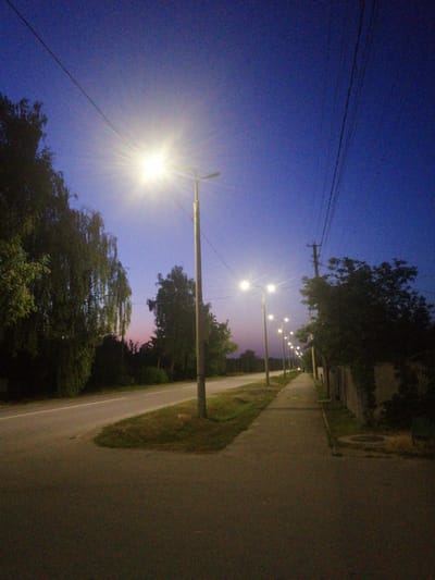 The image depicts a street scene at dusk in Lanivtsi, Ukraine. A paved road runs diagonally from the lower left to the upper right of the frame. A sidewalk runs parallel to the road on the right side of the image. Several streetlights are visible along the sidewalk, illuminating the scene with a bright, white glow. Trees line the left side of the road, some with weeping branches. The sky is a deep blue, with a hint of pink on the horizon, suggesting the last vestiges of sunset. Power lines are strung across the sky. The overall atmosphere is quiet and tranquil, with no people or vehicles visible in the frame. The scene appears to be in a residential or semi-rural area.