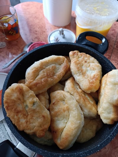 The image shows a black frying pan filled with fried pies. Next to the pan are a glass of tea with a butterfly design, a white sugar bowl with a lid, a spoon, a plastic container of honey, and a roll of paper towels. The text on the side of the pan indicates it is mayonnaise. The text also indicates that the food was created in the Republic of Tatarstan, but does not indicate that the image was taken in Новоалександровка, Russia. The scene appears to be set indoors on a wooden table.
