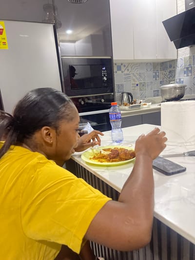 The image captures an indoor scene within a modern kitchen, likely in Adenta Municipality, Ghana, as provided by the location context. The primary subject is a person, appearing to be a woman, with dark hair pulled back, wearing a bright yellow short-sleeved top. She is positioned at a kitchen counter, with her attention directed towards a plate of food.
The activity taking place is eating, or preparing to eat, as a plate containing a meal, possibly rice with chicken or another stewed dish, is directly in front of her on the white countertop. A clear plastic bottle of "PERE" branded water, a black smartphone, and a roll of white paper towels are also on the counter.
Notable details of the setting include sleek black and white kitchen cabinetry. The wall behind the counter is adorned with distinctive blue and white patterned tiles. Visible appliances include a black range hood and a built-in black oven or microwave with a digital display showing blue numbers. To the far left, a silver Hisense refrigerator with a yellow energy efficiency label (partially reading "ENERGY" and "248") is visible. Other kitchen items include a silver pot and a black electric kettle. The lighting suggests an indoor setting, possibly evening or night, illuminated by artificial light. The person's face is partially obscured, so her emotions are not clearly discernible, but she appears focused on her meal.
