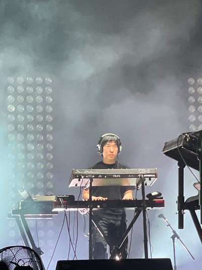 The image appears to show a musician performing on a keyboard synthesizer during a concert. The musician is wearing headphones and dressed in dark clothing. The keyboard is mounted on a stand and surrounded by various pieces of equipment, including a microphone, cables, and a speaker. In the background is a wall of stage lighting that creates a hazy environment. The location may be Salembaran, Indonesia, but this cannot be confirmed from the image itself.