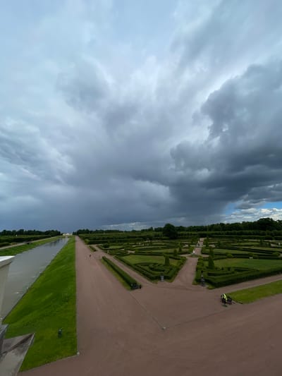 This is a medium-shot, high-angle photograph of a formal garden in Strelna, Russia. The garden features meticulously manicured hedges arranged in geometric patterns, with gravel paths winding through them. To the left of the garden is a long, straight canal with a grassy embankment. In the distance, a light-colored building with a red roof is visible. The sky is filled with dramatic, dark storm clouds, with a small patch of blue sky visible towards the right side of the frame. Two individuals are visible in the garden: one walking on a path and another wearing a bright yellow jacket, appearing to be working or resting on a bench. The overall scene suggests an impending storm in a well-maintained park.