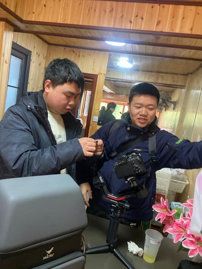 Two Asian men stand inside a wood-paneled room. One man wears a navy jacket and holds a pen, seemingly gesturing. The other wears a similar jacket with a camera and equipment strapped to his chest. A large grey case with the word "HULRC" sits nearby. A table with flowers, a drink, and other items stands in the background. The scene appears to be captured in a slightly disorienting angle, giving the impression of a tilted perspective.