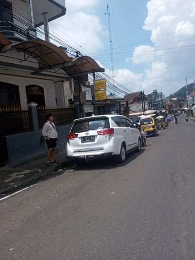 A white Toyota Innova is parked on the side of a road in an urban area. The license plate number is "D 1570 AFA". A man dressed in a white shirt and black shorts is standing near the car on the sidewalk. In the background, there is a yellow taxi van and a variety of other vehicles and people on the street. Buildings line the street, with power lines visible overhead. A minaret can be seen further down the road.