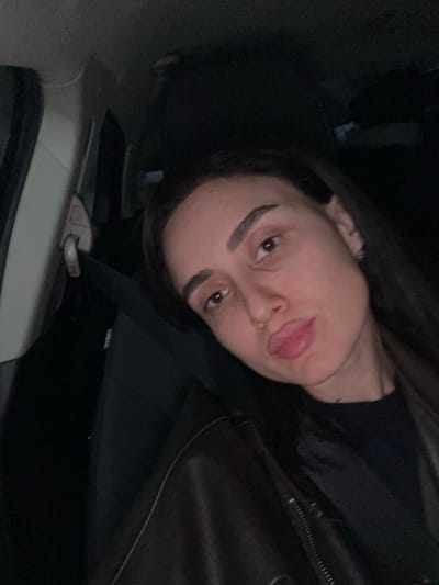 A woman with dark hair is angled and facing the camera with her tongue sticking out. She is wearing a black top and a dark leather jacket. A car seatbelt is visible across her chest. The background includes parts of a car interior, such as a white door panel. Lighting appears to be dim.