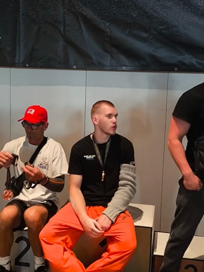 This image captures an indoor scene, likely at a sporting event in Romanshorn, Switzerland, given the presence of medals and podiums. Two men are seated on a podium. The man on the left wears a red cap, glasses, a white t-shirt with "FRANCE" printed on it, and black shorts. He is holding a medal with a bronze finish. The man on the right wears a black t-shirt and bright orange pants, with a grey sleeve on his left arm. A third individual, partially visible on the right edge of the frame, wears a black t-shirt and dark pants, with their arm bent. The background features a dark banner with white speckles, and a light-colored wall. The podium is marked with the number "2" on the left side.
