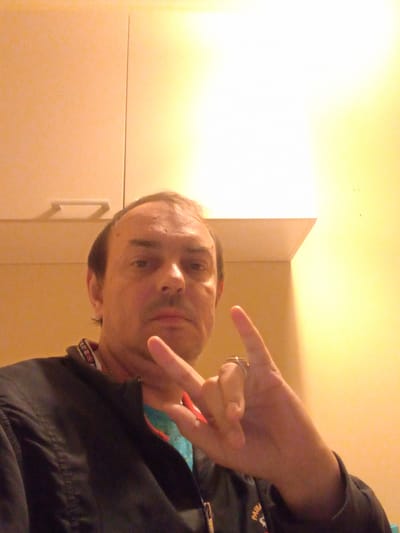 A Caucasian male, appearing to be in his 40s, is captured in a selfie-style photograph. He has short, dark hair, a visible hairline, and appears to be clean-shaven or with light stubble. His expression is neutral. He is wearing a black jacket with a collar that has a repeating pattern of text and symbols. Beneath the jacket, a blue garment is partially visible.

The man is positioned in what appears to be an indoor setting. Behind him, a light-colored wall is visible, with a horizontal line suggesting cabinetry or a shelf. To the right, the wall transitions to a bright yellow hue, illuminated by an overhead light source. The lighting casts a distinct shadow and a bright, blown-out area on the wall.

The man's right hand is raised, with his index and pinky fingers extended, forming a 'rock on' gesture. A ring is visible on his right ring finger. His left hand is not clearly visible. The overall image has a slightly grainy quality, possibly due to low light conditions or camera settings.

There are no explicit visual cues in the image that indicate the location is within the city of Kajaani, Finland. The provided environment could be a residence, an office, or another indoor space.