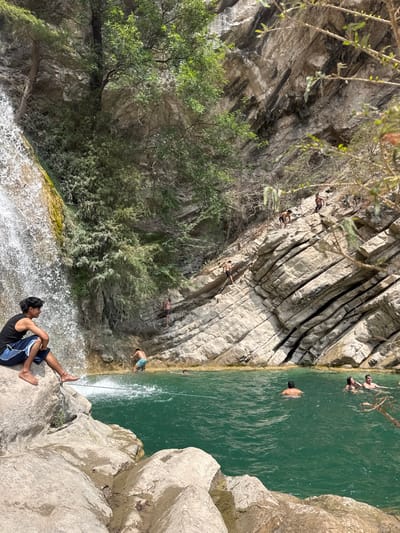 This image depicts a natural swimming area located in San Miguel Atlapulco, Mexico, under bright daylight conditions. A large waterfall cascades from the upper left into a clear, teal-colored natural pool that occupies the lower half of the frame. The left side of the scene features the waterfall, its base surrounded by large, light-colored rocks. Dense green vegetation and trees cling to the rocky cliffs above and to the left of the waterfall.
The right side of the image is dominated by steep, layered rocky cliffs, showing distinct horizontal stratification. Scattered vegetation, including some dry brush, grows on these cliffs.
Approximately ten individuals are present. In the immediate foreground, a young male with dark hair, wearing a black tank top and blue and white shorts, sits barefoot on a large, light-colored rock. He has an ankle bracelet on his left ankle and is looking towards the right side of the pool. His feet are positioned near the water's edge, close to a dark rope that extends across the water towards the right.
Within the pool, several individuals are swimming or wading. One male in light-colored shorts stands in the shallow water near the base of the waterfall, facing away from the camera and creating a splash. Further into the pool, at least four other individuals are visible; one male with a bare back is submerged to his shoulders, and two individuals (one male and one female) are positioned closer to the far bank, appearing to interact. Another person's head is partially visible in the water.
On the steep rocky cliff face to the right, multiple individuals are engaged in climbing or descending. Three males are distinctly visible on the rock layers: one higher up, wearing dark shorts; another below him, also in dark shorts; and a third further down, appearing to use a rope for assistance or support. Another individual is visible near the water's edge at the base of the cliff. The rope seen spanning the water in the foreground also connects to the cliff face on the right, and another rope extends down the cliff face towards the water.