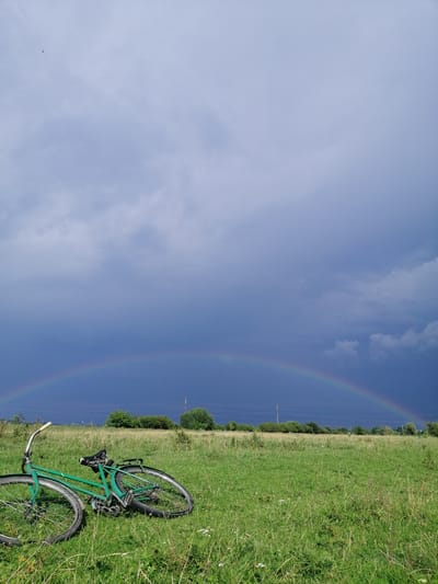 In a grassy field, a green bicycle lies on its side. The bicycle is positioned in the foreground, with its wheels and frame clearly visible. Beyond the field, a line of trees forms the horizon, with utility poles scattered amongst them. The sky above is dramatic, with dark, stormy clouds dominating the upper half of the image. A faint rainbow arcs across the sky, a colorful contrast to the brooding clouds. The overall scene suggests a post-storm landscape in the city of Krasnoluka, Ukraine.
