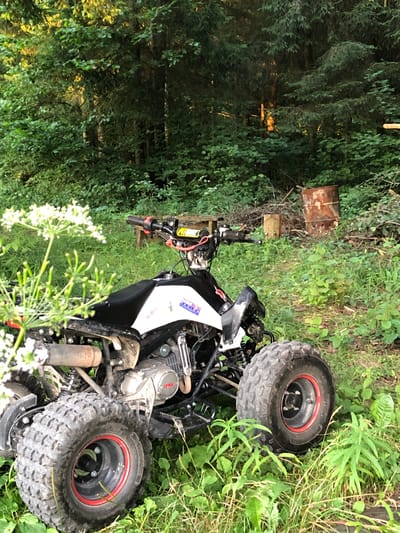 An all-terrain vehicle (ATV) is parked in a grassy, wooded area. The ATV is black and white with red accents on its wheels. It has large, knobby tires. The background consists of dense green foliage, with sunlight filtering through the trees. There are some discarded items in the background, including a rusted barrel and some wooden planks. The overall scene suggests a rural or semi-rural setting.