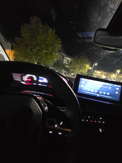 An interior car view at night shows a brightly lit touch screen display against the dark dashboard. A portion of the steering wheel is visible, with illuminated buttons for calls, source, and list options. Through the windshield, streetlights and trees are visible. A rearview mirror is also present in the upper left corner. The scene suggests the driver is either parked or slowly moving through a residential area.