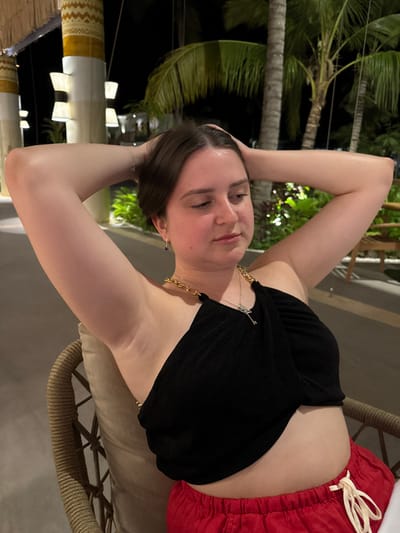 A woman poses while sitting on a chair. She wears a black top with gold chain straps and red shorts with a white drawstring. Her hair is short and dark. She has both arms raised behind her head. Palm trees are visible in the background. The photo appears to be taken outdoors at night.