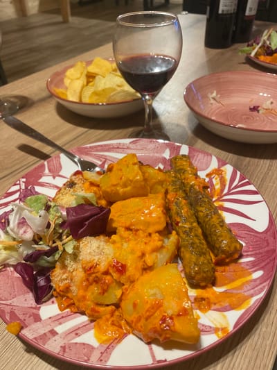 The scene depicts a meal served on a wooden table in an indoor setting in the city of Palež, Montenegro. In the foreground, a white plate with a red/pink leaf pattern holds a main course consisting of chunks of potatoes coated in a thick, bright orange-red sauce, two cylindrical meat items also covered in the same sauce, and a side portion of mixed green salad with shredded vegetables. A silver fork rests on the plate. To the left, a white bowl with a red rim contains plain potato chips. Centered in the midground is a wine glass containing red wine, approximately one-quarter full. To the right of the wine glass, an empty white bowl with a red rim and leaf pattern, matching the main plate, contains minimal salad remnants. In the blurry background, dark-colored bottles and light-toned furniture are faintly visible. No people are present.