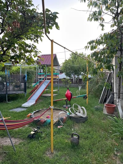 This image depicts an outdoor backyard scene in Pîrlița, Moldova, likely on a cloudy day. The yard is filled with various objects, suggesting a space used for recreation and storage. In the foreground, a yellow horizontal bar, possibly part of a swing set or play structure, stretches across the frame. To the right of this bar, a red punching bag hangs, indicating a workout area. Further to the right, a large black kettlebell sits on the grass, and a tire with some white circular objects is also present.
In the midground, a colorful slide with a metal surface is set up, with a wooden frame supporting it. Behind the slide, a house with a burgundy roof is visible, partially obscured by trees and foliage. There's a metal fence in the background.
The yard is covered in green grass, with some areas showing bare earth or gravel. Several trees with green leaves are scattered throughout the scene, with one prominent tree on the left side of the image. In the far background, more houses and trees can be seen, typical of a village or suburban setting.
There are no people visible in the image, and the overall atmosphere seems quiet and still. The presence of play equipment and weights suggests this is a private backyard, likely belonging to a family. The objects scattered around, such as the tires and a terracotta pot on the left, might indicate a need for tidying up or a deliberate rustic aesthetic. The lighting suggests it is daytime, but the overcast sky prevents strong shadows.