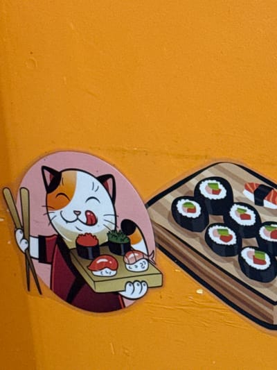 A close-up shot captures two cartoon stickers affixed to an orange surface. The stickers feature sushi themes. The upper sticker displays a wooden tray filled with an assortment of sushi rolls, including maki with seaweed exteriors and fillings of cucumber, salmon, and other ingredients. Below this, a round sticker depicts a cartoon cat with a cream-colored face, orange markings, and closed eyes, holding a sushi tray. The cat is dressed in a red robe with black accents, and chopsticks lie at its feet. The location, Novo-Nikol'skoe, Russia, cannot be confirmed from the image alone.
