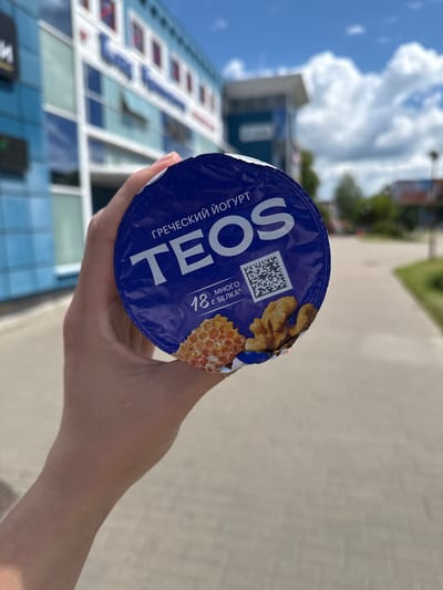A person's hand holds a blue yogurt cup with white text and graphics. The yogurt cup is oriented diagonally, with the person's thumb and fingers curled around its side. The visible text on the cup includes "TEOS" in large white letters, "ГРЕЧЕСКИЙ ЙОГУРТ" in smaller white text below "TEOS", and some smaller text in Russian that appears to say "18 г. БЕЛОК" which translates to "18 g. PROTEIN". There is also a QR code and graphics of walnuts and honeycomb on the cup.

The background is blurred, suggesting the image was taken with a shallow depth of field. Visible elements in the background include a modern building with blue and white windows, a sidewalk or paved pathway, and a cloudy sky with some blue patches. The lighting suggests it is daytime. The setting appears to be an outdoor urban environment. The image conveys a casual, everyday moment, possibly someone holding a snack outdoors.
