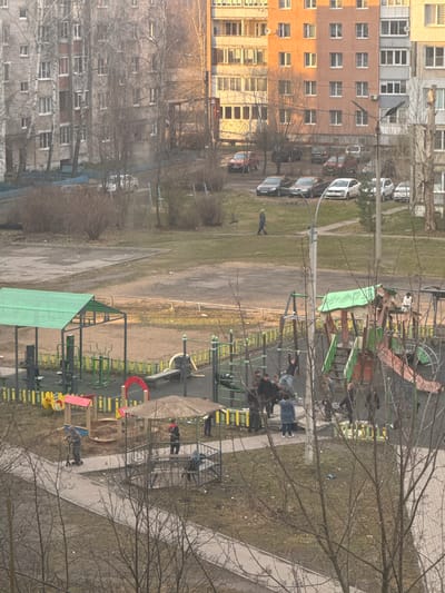 An elevated, oblique-angle view depicts a residential area in Kladovitsy, Russia, with apartment buildings to the left. A playground is prominent in the foreground and midground, featuring slides, climbing structures, and covered play areas. Bare trees are interspersed throughout the scene. Several individuals, including adults and children, are present on the playground. Some are standing and interacting near the climbing structures, while others are dispersed throughout the area. Parked cars are visible on a street between the playground and the apartment buildings. The environment suggests a late fall or early spring setting.