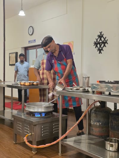In Perumannur, India, three men are in a kitchen area, possibly a cooking demonstration or class. The man in the foreground, wearing a purple shirt and a red and blue checkered apron, is actively pouring something from a stainless steel container into a large pot that is heated by a blue gas flame on a stainless steel stove. To his left, another man, wearing a light blue shirt and a checkered apron, stands by a metal table with several small copper bowls. Further left, a third man in a light blue shirt observes the cooking process. The environment features stainless steel countertops, gas cylinders, and wall decor, including a clock and an abstract design.