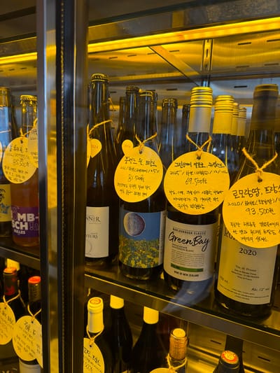 The image shows a shelf in a refrigerated display case filled with wine bottles. The location appears to be a store or restaurant in Toseong-myeon, South Korea, given the presence of price tags with Korean text and currency symbols. Several bottles have yellow circular tags attached with descriptions and prices written in Korean.

Notable wine brands visible include "Green Bay" Marlborough Classic Sauvignon Blanc from New Zealand and "Vin de France" from France. Other bottles display labels in French, such as "ALSACE VEEURS".

The bottles are densely packed, and the lighting within the case is predominantly yellow. The display shelves are made of a reflective metal material. The focus is sharp on the central bottles with the tags, while those in the foreground and background are slightly out of focus.