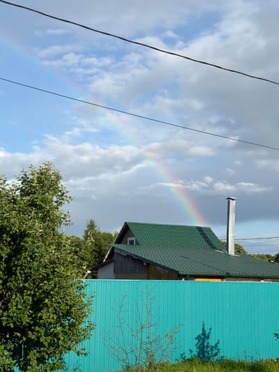 A wide shot captures a suburban scene in Satino-Russkoe, Russia, under a partially cloudy sky where a faint rainbow arcs across the upper left quadrant. In the foreground, a vibrant turquoise wooden fence stretches horizontally, partially obscuring the view of a residential house with a dark green metal roof. A tall, silver metal chimney stands on the roof, to the right of a small attic window. To the left of the house, a lush green tree with dense foliage dominates the frame, its branches reaching towards the sky. Beyond the house, a line of trees creates a natural backdrop, with a visible power line running across the top of the image. The ground in front of the fence is covered in green grass, and shadows are cast upon the fence from unseen objects. There are no people or animals visible in the frame.