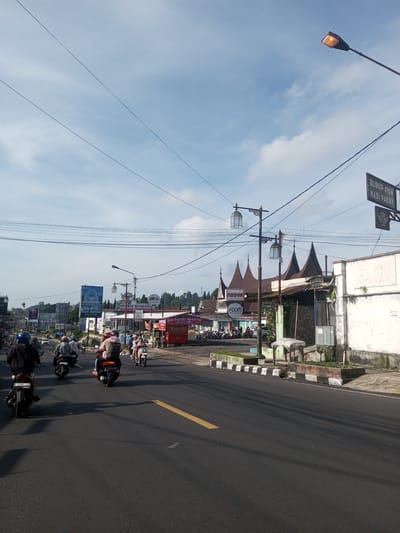 The image depicts a street scene in a Southeast Asian town. Motorbikes occupy the main street. The sky is partly cloudy. Along the roadside are restaurants and shops, including one with distinctive Minangkabau-style roofs. Signage advertises "RM Putri Minang" (a Padang restaurant) and "Bubur Ayam Nasi Paket" (chicken porridge with rice set). Power lines stretch across the sky. A yellow line divides the asphalt road. Vegetation and concrete barriers border the street.