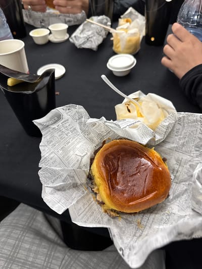 The image shows a hamburger and fries served in newspaper-printed wrapping on what appears to be a black surface. Partially visible are a person's arm and hand, a water bottle, and several small white cups, possibly containing condiments. The setting appears to be indoors, possibly a restaurant. San Bartolomé, Spain, is not discernible from the details present in the image.