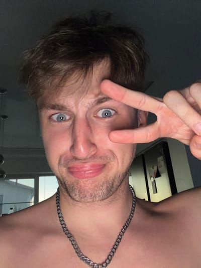 A close-up, selfie-style photograph shows a young Caucasian man with light brown, tousled hair and light blue eyes. He is looking directly at the camera with a playful expression, his lips pursed and his left eye covered by his right index and middle fingers forming a peace sign. He is wearing a thick, silver chain around his neck. The background is dimly lit and out of focus, showing what appears to be an indoor setting with a window on the left side of the frame and a framed picture or poster on the right. There is no indication or visual evidence connecting this image to the city of Vanino, Russia, or any specific urban environment.