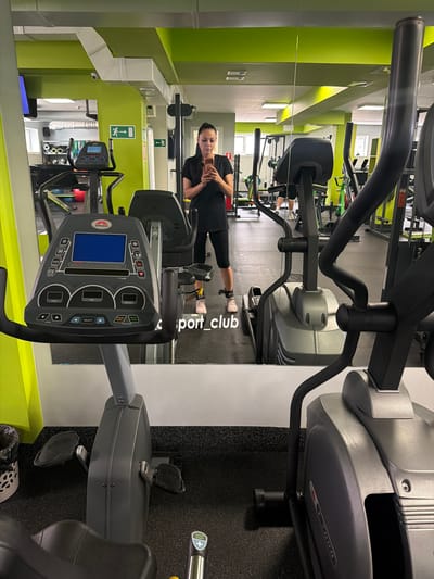 A woman is taking a mirror selfie in a gym. She is wearing a black t-shirt and black leggings, with pink sneakers and ankle weights. The gym appears to be well-equipped with various exercise machines, including stationary bikes and elliptical trainers. The walls are painted in bright green, and the floor is covered with black rubber mats. The image is taken from the perspective of the woman, looking into a large mirror that reflects the entire gym. In the background, other gym-goers can be seen exercising on different machines. The overall impression is that of a modern and active fitness center. The gym is located in Timashevsk, Russia.