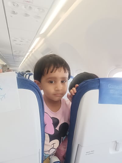 A young child with dark hair and brown eyes is featured prominently in the foreground, peeking over the backrest of an airplane seat. The child, likely between 3 and 5 years old, wears a light pink t-shirt with a Minnie Mouse graphic and looks directly towards the camera with a neutral, slightly inquisitive expression. Their hands are resting on the blue fabric covering the top of the seat back.
The setting is the interior of an airplane cabin, visible through bright overhead lighting and natural light from a window on the right. The cabin features rows of blue and white airline seats. The seat backs are primarily white plastic, with a blue patterned fabric on the upper sides and headrest area. Overhead panels with individual air vents and reading lights are visible towards the ceiling.
Behind the child, the dark hair of another passenger in the seat in front is partially visible. On the right side, the back of an airplane seat shows a blue headrest cover with partially visible white text that appears to read "Comfor" and "stayfor", suggestive of an airline branding (likely "Comfort Zone" or similar). Below this, on the white plastic seat back, black text reads "धूम्रपान निषेध" (Dhumrapan Nishedh - Hindi for "No Smoking") followed by "NO SMOKING" in English. Another "No Smoking" symbol (a crossed-out cigarette) is visible on an overhead panel.
The scene captures a moment during an air journey, possibly during boarding or mid-flight. The bright lighting suggests it is daytime. The blue and white colors of the seats, coupled with the "NO SMOKING" signage in both English and Hindi, indicate a commercial airline, potentially an Indian carrier given the location context of Anneshvara, India.