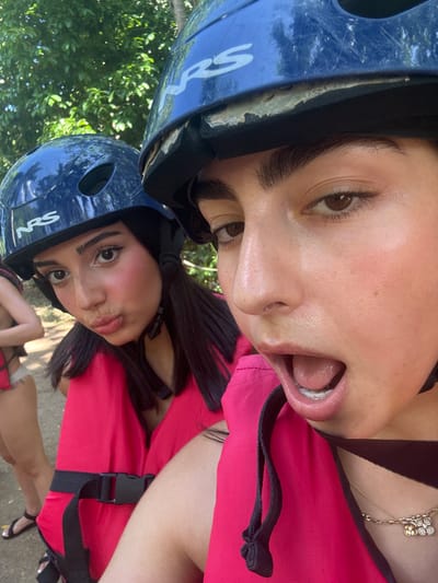 The image captures three light-skinned females outdoors in Woods Town, Jamaica. The foreground features a female with dark hair, wearing a blue NRS brand helmet with visible wear and a bright pink personal flotation device. Her mouth is open wide, and her eyes are directed at the camera. A delicate gold necklace with a multi-part pendant is visible. To her left, another female with dark hair wears an identical blue NRS helmet and a bright pink personal flotation device with a black buckle; she presents a "duck face" expression towards the camera. In the far left background, a third female, partially visible, stands on a dirt path, wearing a light-colored bikini bottom, dark sandals, and a reddish garment. The background consists of dense green foliage under bright daylight. The attire and equipment suggest preparation for an outdoor or water-based activity.