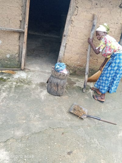 The image captures an outdoor scene in Kamantan, Nigeria, on October 25, 2012, at 06:48 AM. An elderly woman, wearing a yellow floral top, a blue and white patterned long skirt, black sandals, and a white and olive-green headscarf, is depicted actively sweeping a concrete ground surface. She holds a natural fiber broom in her right hand and a long wooden pole, possibly for support, in her left hand, leaning forward as she sweeps. To her left, a dark, cylindrical wooden mortar or stool is present, topped with a folded light blue and pink patterned fabric. Behind these objects, a doorway with a wooden frame leads into a dark interior of an earth-built structure. A square-nosed shovel, partially covered with swept organic debris, lies on the concrete ground in the foreground. A small quantity of additional natural fibers or debris is situated near the doorway's left side. The building's wall exhibits an earth-toned, textured finish.