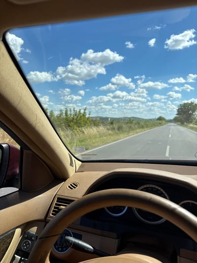 The image shows a point-of-view perspective from the driver's seat of a car on a sunny day. The road ahead is visible, stretching into the distance with a dotted white line marking the lanes. The sky is blue with scattered white clouds. The car's interior, including the steering wheel, dashboard, and a portion of the side mirror, is also visible in the foreground. The surrounding environment appears to be rural, with dry grass and some trees lining the road. The dashboard indicates the car is traveling at approximately 70 km/h. There are no people or other vehicles visible in the frame. The metadata suggests the image was captured in Sotirya, Bulgaria.