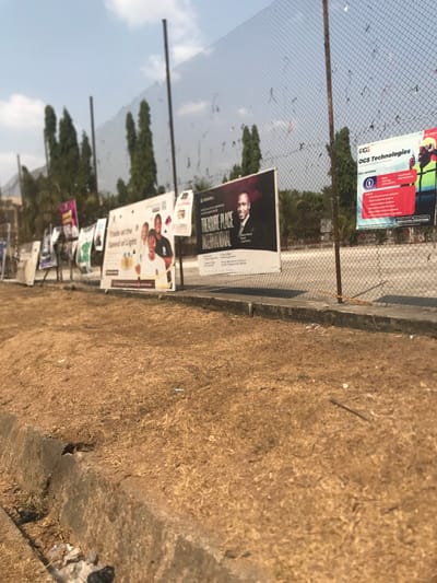 The image captures an outdoor scene in Umudike, Nigeria, during what appears to be a bright, sunny daytime under a clear blue sky with some scattered clouds.
A wire mesh fence runs diagonally across the mid-ground, serving as a display for numerous advertisements and posters. In the foreground, a strip of dry, light brown grass and dirt occupies the lower right section, bordered by a concrete curb or edge to its left.
Several large billboards are prominently attached to the fence. The top-most legible one is red and green, advertising "OGS Technologies" and listing services such as "CCTV," "Networking," "Software Development," "Web Design & Hosting," "Training," and "Sales & Maintenance."
Below this, a significant billboard with a black background features a portrait of a bald man in a suit. Text on this poster reads "THE TREASURE PLACE INTERNATIONAL," followed by details like "TPW Global," "Prayer & Worship," "Every Thursday," "5:00 PM," and a specific location: "RCCG Strong Tower, Umuahia - Aba Rd, Beside Ubakala Market, Umuahia, Abia State, Nigeria."
Further along the fence, another noticeable billboard with a white background displays several people and advertises "Trade at the Speed of Light," with partial mentions of "Premier League" and "Fantasy Football." Numerous other smaller and indistinct posters are also visible further down the fence line.
Behind the fence, a line of green trees is visible, separating the fence from the clear sky. No specific human activity is depicted, but the prevalence of advertisements suggests commercial promotion in a public area.