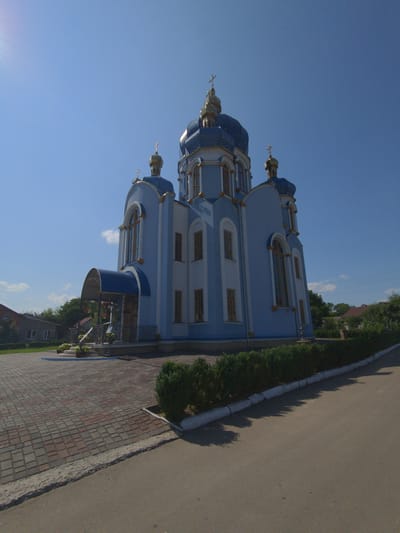 The image depicts a large, light blue church with a blue, golden-domed roof and golden crosses. The church has several windows with arched tops and golden trim. In front of the church is a paved walkway made of red and gray bricks, leading up to an entrance with a blue awning and railings. To the left of the church, there are some houses and trees. In front of the church, there is a neatly trimmed green hedge running along the edge of the paved area, and beyond that, a paved road. The sky is clear blue with a few small white clouds. The sun is shining brightly, casting shadows. The scene appears to be a sunny day in a quiet, possibly rural or suburban, area. The church is the central focus of the image, suggesting it is a significant building in the community. This church is located in the city of Zalishchyky, Ukraine.