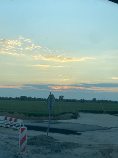 The image captures a scene at a construction site in Podłęże, Poland, during what appears to be either sunrise or sunset, given the soft, diffused light and colored clouds in the sky. The foreground is dominated by construction materials and temporary barriers, including a white and red striped barricade and a similar-looking post. There is disturbed earth and what looks like unfinished road or paving.
In the middle ground, a muted gray, octagonal traffic sign is visible on a pole, its purpose unclear without further context. Beyond this, a vast, green agricultural field stretches out, bordered by a line of trees and vegetation in the far distance. The sky is a prominent feature, transitioning from a pale blue at the top to hues of yellow, orange, and soft pink near the horizon, with scattered wispy clouds. Faint contrails from aircraft are visible in the upper sky. The overall impression is one of a rural or semi-rural location undergoing development, with the natural beauty of the sky providing a backdrop to the human activity.