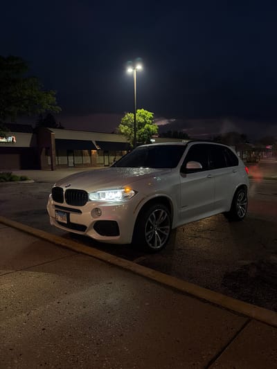 A white BMW X5 is parked at night on a wet asphalt lot in front of a strip mall. The vehicle's headlights and taillights are illuminated. The sky is dark with some clouds visible. A street light illuminates the scene. The license plate reads "EV 79644". The location is Buffalo Grove, United States of America.