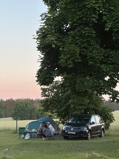This image depicts a campsite scene under a large tree, likely located in Завалочыцкі сельскі Савет, Belarus, given the license plate of the car. In the foreground, a black Volkswagen Sharan minivan is parked on the grass. To the left of the car and partially under the tree's canopy, a green tent is set up. A portable table is placed in front of the tent, laden with items such as plates and bottles. Four people are seated around the table, appearing to be engaged in conversation and enjoying a meal or drinks. Two individuals are sitting on folding chairs, while two others are seated on the ground or directly on a mat. The background consists of a grassy field with a fence, and a dense forest can be seen in the distance. The sky exhibits a soft, pastel gradient, transitioning from light blue at the top to pinkish hues near the horizon, suggesting it is either dawn or dusk. The overall atmosphere is one of a relaxed outdoor gathering.