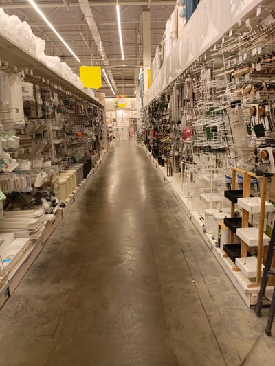 The image depicts the interior of a large retail store, likely a home goods or hardware store, with aisles of merchandise on either side and a concrete floor. The perspective is from within one of the aisles, looking down the length of the store. The shelves are filled with various items, including kitchenware, storage containers, and cleaning supplies. The lighting appears to be artificial, likely from fluorescent or LED fixtures on the ceiling, which is not fully visible but shows structural elements and ductwork. There are no people visible in the image. The store environment appears organized and well-stocked, typical of a commercial retail space. Based on the provided crop hints and the context of "Дядьково, Russia," this scene is a retail store located in Дядьково, Russia.