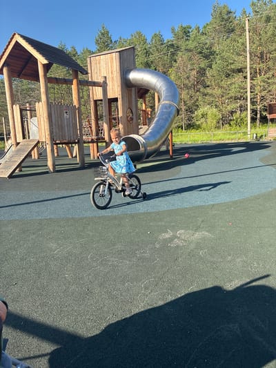 A young girl in a blue dress with a unicorn pattern rides a white bicycle with a black basket. She is on a playground with a large wooden structure featuring a spiral slide. The playground surface is a combination of dark green and blue rubber. In the background, there are tall pine trees and a bright blue sky. A red ball is visible on the ground near the slide.