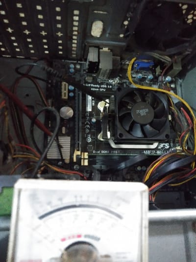 The image displays a detailed, forensic view of the interior of a desktop computer case, located in Mornington, Australia. The primary subject is a dark-colored motherboard, identified as model A68F2P-M4 V1.4. A black CPU cooling fan, labeled "ZPJ6018281 5615", is centrally mounted on the motherboard's heatsink. Legible text on the motherboard includes "G.P. Ready USB 3.0", "32nm CPU", and "Dual DDR3 2133". A blue VGA port is visible towards the upper right. A dense array of multicolored cables, including red, yellow, black, and orange, are connected to various components within the case. The upper portion of the computer case, featuring a perforated metal grill, is visible at the top of the frame. In the out-of-focus foreground, the upper half of an analog multimeter's dial and needle are present, indicating potential diagnostic activity. No individuals are depicted.