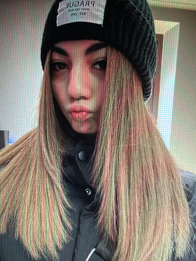 A young woman with long, blonde hair and a black beanie is captured in a close-up, head-and-shoulders selfie. She is wearing a dark jacket. The background appears to be an indoor setting with a wall featuring a subtle grid pattern and a vertical light source. There are no visible indications of the location in Chotěšov, Czechia.