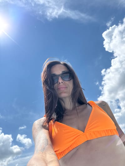 A woman wearing an orange bikini and sunglasses poses for a low-angle selfie against a bright blue sky with scattered white clouds. Sunlight glints off her sunglasses and illuminates her face and chest. Her dark hair is windswept. She has tattoos on her left shoulder and arm. The image is captured from a perspective looking up at the woman, emphasizing the expanse of the sky above. The location is specified as Grivitsa, Bulgaria.