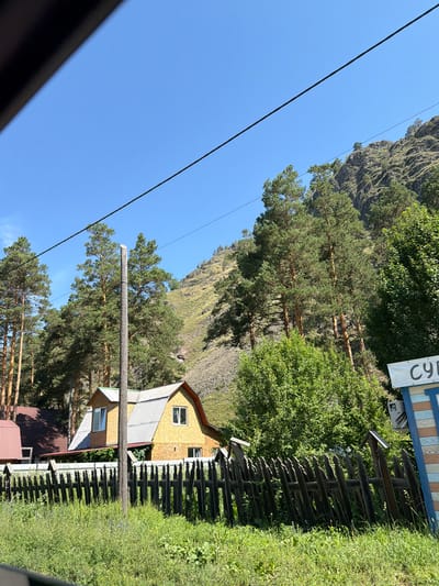 The image depicts a rural scene with a house and a fence in the foreground, set against a backdrop of a steep, rocky hillside covered in pine trees. The house is a two-story structure with a yellow exterior and a corrugated metal roof. A dark wooden fence with pointed pickets runs across the front of the property. Tall pine trees are abundant, with some visible on the lower slopes of the hill. The sky is clear and blue. A white sign with blue lettering is partially visible on the right side of the image. The scene appears to be an outdoor environment in a natural setting.