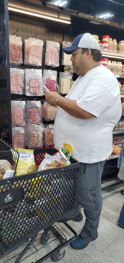 The image captures a male shopper in the deli section of a supermarket, likely in Barquisimeto, Venezuela. The man, depicted from the waist up and slightly to the right, is wearing a white short-sleeved t-shirt, gray sweatpants with "Reebok" written on the left leg, a blue and white baseball cap, and blue sneakers with yellow accents. He has short dark hair and a beard or heavy stubble. On his right wrist, he wears a beaded bracelet featuring a silver cross charm.

He is bent forward, appearing to select an item from a brightly lit refrigerated display case that holds numerous vacuum-sealed packages of various deli meats, including ham, salami, and bacon, as well as slices of cheese. His right hand is reaching towards a package of red, circular deli meat.

To the right, a black, plastic-coated metal shopping cart is partially visible. It contains several items, including a yellow package labeled "Natural Mix" cereal and other foil-wrapped products. The cart also has a small white label on its handle area that reads "RIO Supermarket" with a logo. The supermarket floor is light-colored and tiled. The overall scene suggests a routine grocery shopping trip during daylight hours, given the bright indoor lighting.