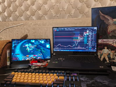 The image shows a desk setup with two laptops, a keyboard, and various other items. One laptop displays a video game interface, likely World of Warcraft, with character information and a game map. The second laptop shows a cryptocurrency trading chart with price data and technical indicators. A yellow and black mechanical keyboard is in the foreground, with RGB backlighting. Other objects on the desk include a tube of lip balm, cables, and a small painting or print depicting figures in front of a cityscape. The background features textured wallpaper and a patterned border. There is no discernible information within the image that would allow for determination of its location in the city of Rubtsovsk, Russia.