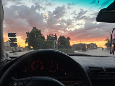 The image is taken from inside a car, looking through the windshield. The dashboard with its illuminated gauges is visible in the foreground. The sky outside is filled with dramatic clouds, colored by a sunset with hues of orange, pink, and turquoise. Through the windshield, a street scene is visible in the distance, depicting buildings, trees, a statue, streetlights, and several people walking. The scene appears to be a town or city in Bulgaria, possibly Letnitsa, based on the mention of the city. The overall atmosphere is that of a warm evening drive.