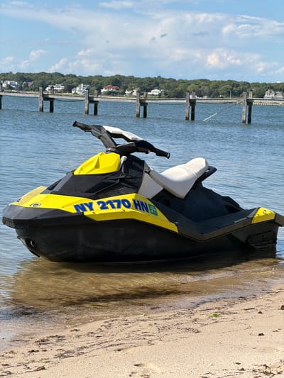 A yellow and black jet ski with the registration number "NY 2170 HN" and a small green sticker with the number "27" is partially submerged in shallow water on a sandy beach. The jet ski is positioned on the right side of the frame. In the background, there is a wooden pier extending into the water, with several pilings. Beyond the pier, a treeline of green foliage is visible, with houses nestled amongst the trees. The sky is blue with scattered white clouds. The scene appears to be located in Hampton Bays, United States of America.